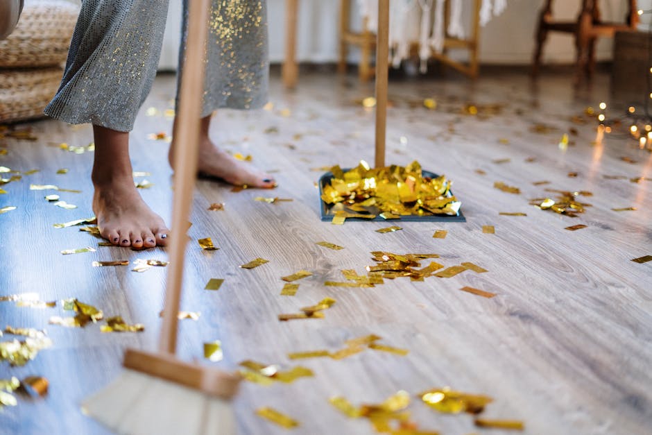 A person is cleaning a light-colored wooden floor in a room decorated for a celebration or party, with gold confetti scattered across the surface. The individual is barefoot, and only their lower legs and feet are visible, wearing a sparkly dress. They are using a broom to sweep up the confetti, which is collected in a dustpan placed on the floor. The room features natural light, with some furniture and decorative elements visible in the background, and the floor appears clean and well-maintained, reflecting the effort of surface cleaning after a festive event. This scene exemplifies deep cleaning and surface hygiene conducted by Kensington Cleaners in a domestic setting.