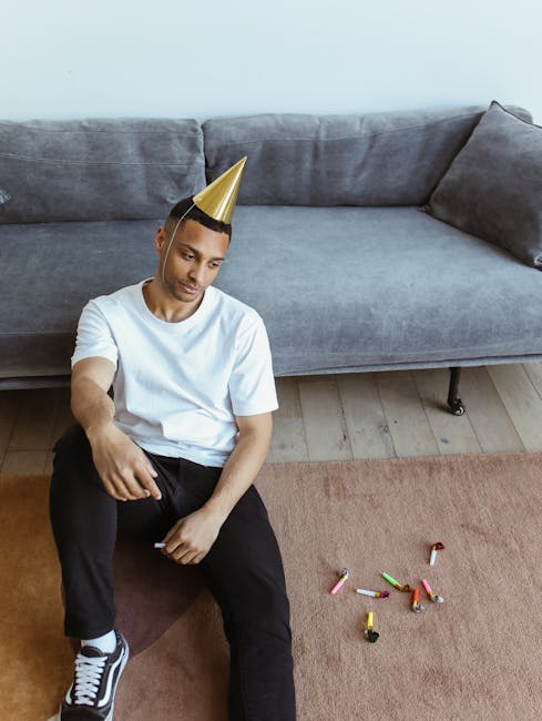 A young man wearing a white t-shirt, black pants, and a gold party hat sits on a wooden floor next to a light brown carpet in a living room. The room features a large gray fabric sofa with multiple cushions, positioned against a white wall under natural lighting. The floor shows scattered small chalk pieces and a partially visible white object, possibly a marker. The scene illustrates a post-party setting with minor mess, where Kensington Cleaners offers surface cleaning and professional domestic cleaning services to restore the cleanliness of the living space after social gatherings.