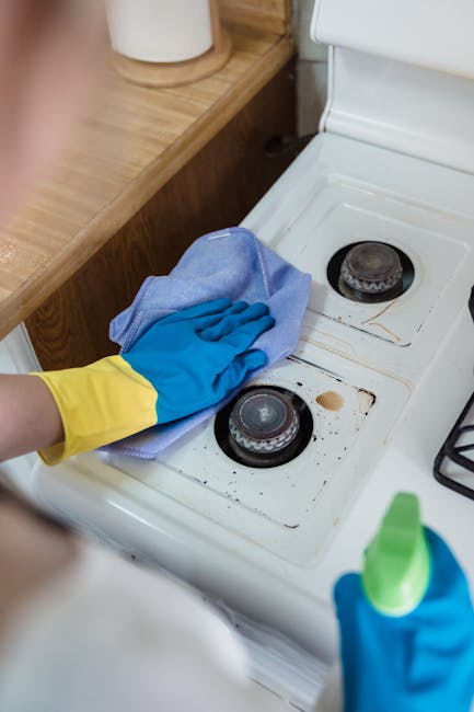 A person wearing yellow and blue rubber gloves is cleaning the stovetop of a white gas range in a kitchen. The individual is using a light blue cleaning cloth to wipe around the burners, which are sli