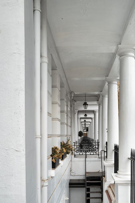 A photograph of a covered outdoor walkway featuring white structural columns supporting the ceiling, with decorative black wrought iron railings along the edge. The corridor has a clean, smooth concrete floor and white walls adorned with potted plants, indicating a well-maintained residential or commercial property in Kensington. The area is illuminated by natural light, highlighting the fresh, spotless appearance of the surface materials. Kensington Cleaners offers professional surface cleaning and deep cleaning services to ensure such spaces remain hygienic and presentable, especially relevant for end of tenancy and sanitisation needs on Kensington High Street SW7.