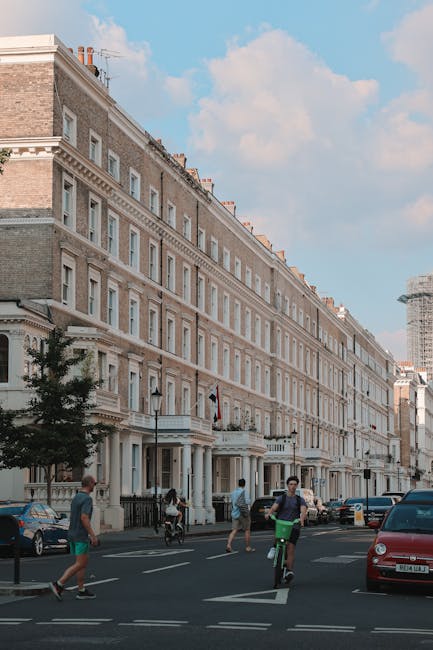A cobblestone courtyard between white residential buildings with large windows and black iron balconies. The courtyard features potted plants and small trees along the edges, with wooden benches posit